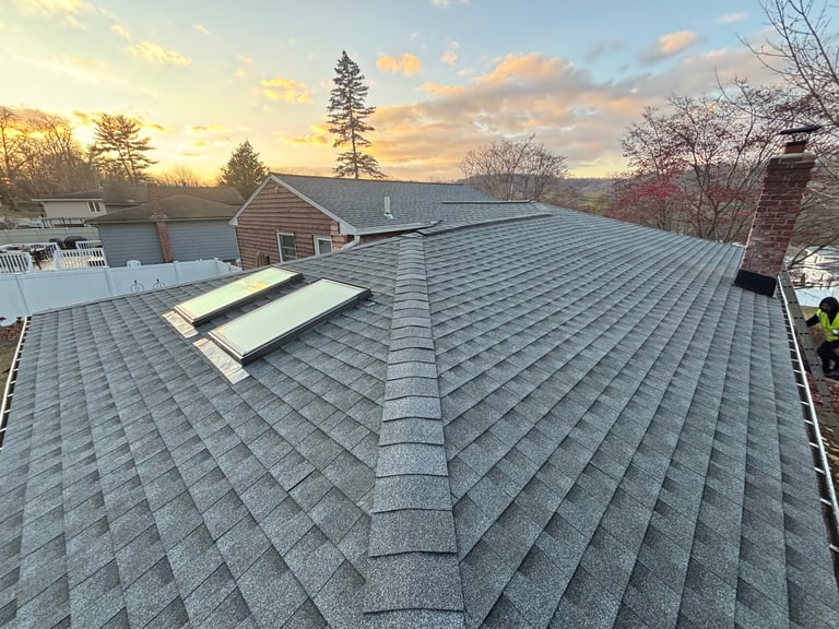 Aerial view of a residential roof with asphalt shingles, ridge vent, and skylights overlooking a lake at sunset