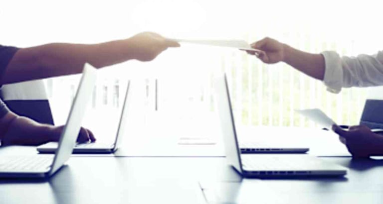 Two people exchanging a document across a desk with laptops and notebooks in a business meeting