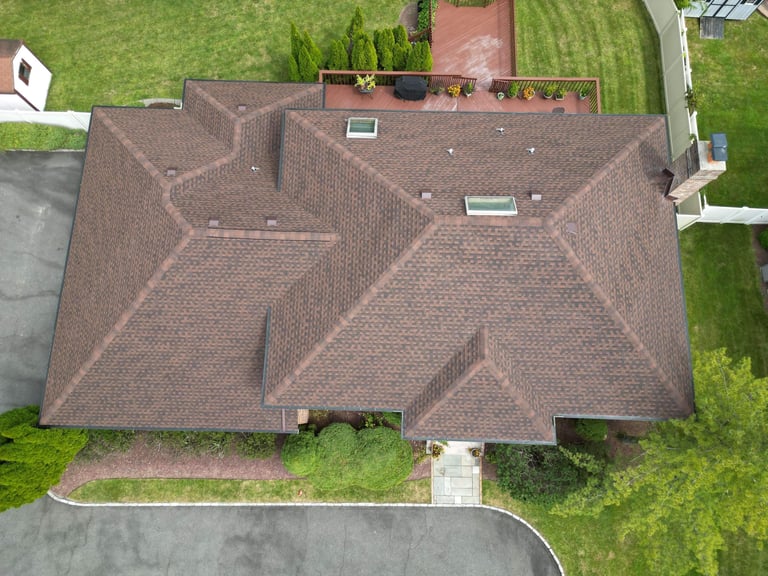 Aerial view of a residential house with brown shingled roof, surrounded by green lawn and driveway