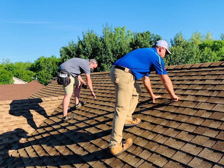 Two workers in safety gear inspect and work on a residential asphalt shingle roof on a sunny day